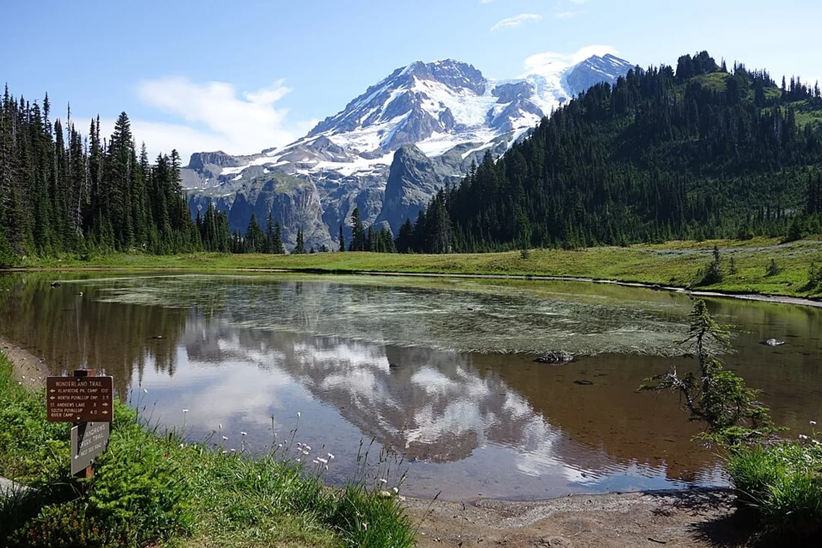 A still mountain lake reflects a view of Mount Rainier. A trail sign is planted on the lake's shore. Aurora Lake is a popular landmark in Klapatche Park along the Wonderland Trail, with views of Mount Rainier. Keywords: Mount Rainier; trails; hiking; Klapatche Park